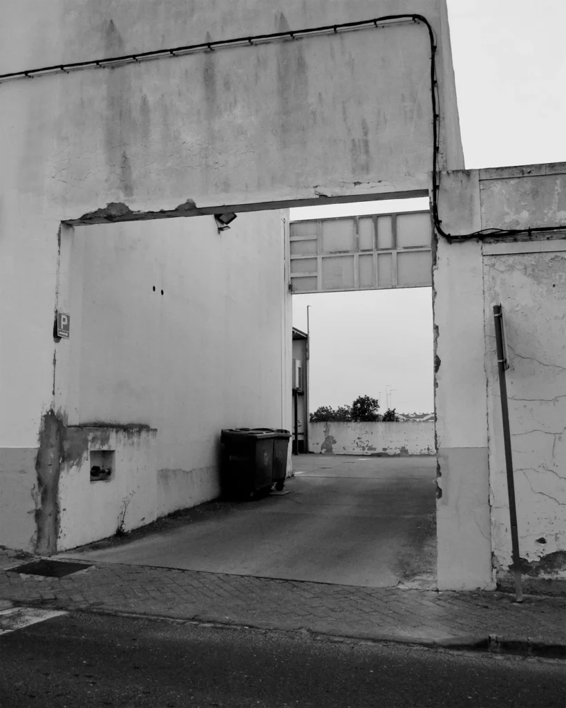 Black and white photo of a weathered whitewashed archway in a Portuguese town, with cracked plaster, a cable running along the wall, and a quiet courtyard beyond.