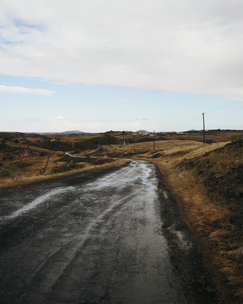 Wet, cracked asphalt road winding through dry golden hills in the Alentejo, with power lines leading toward a white village on the horizon under a cloudy sky.