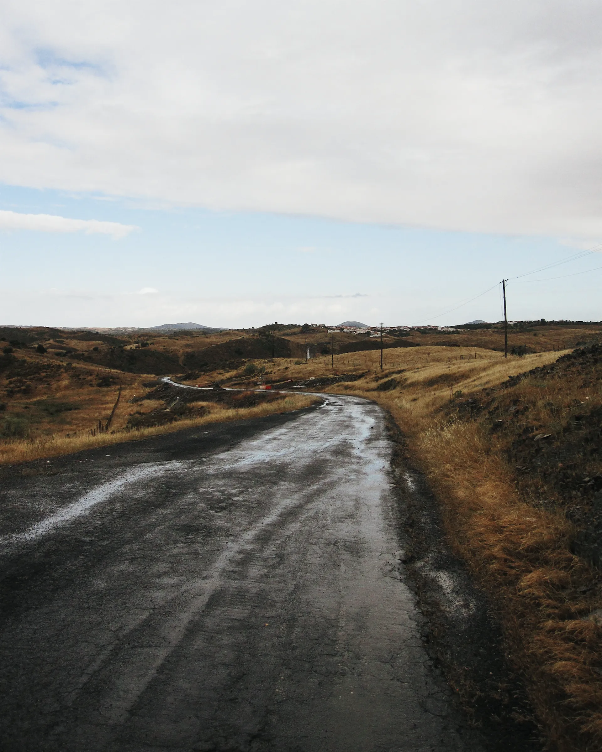 Wet, cracked asphalt road winding through dry golden hills in the Alentejo, with power lines leading toward a white village on the horizon under a cloudy sky.