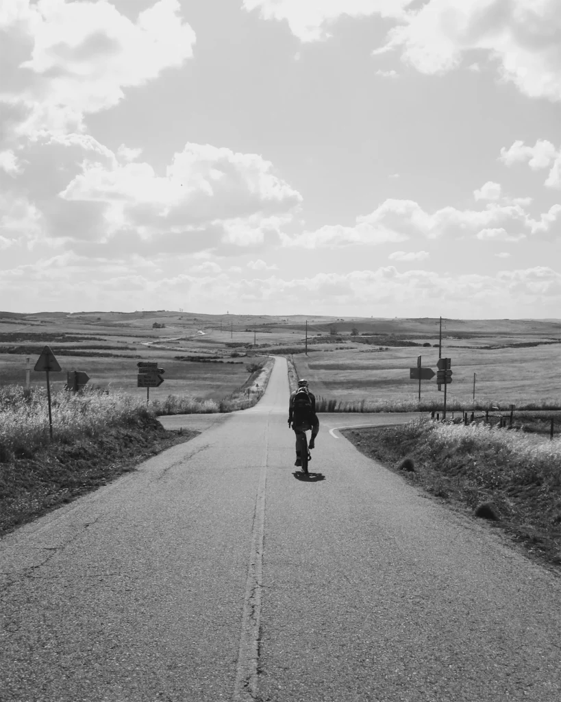 Black and white photo of a cyclist riding away down a long straight road through open rolling plains, passing a crossroads with signs to Castro Verde and Beja.