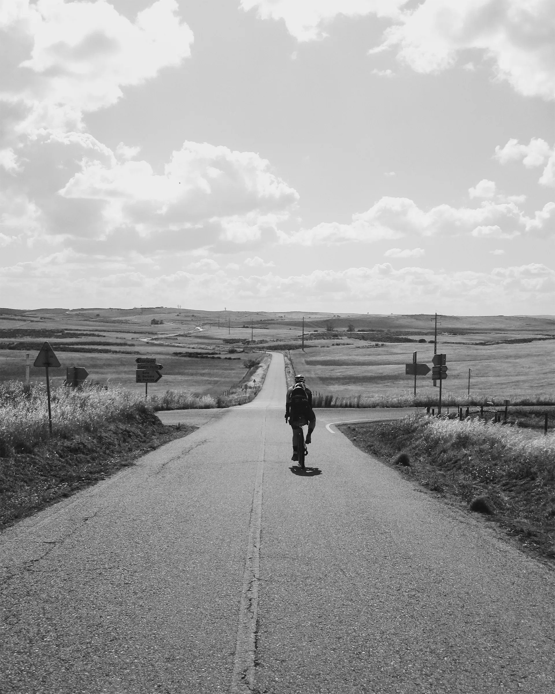 Black and white photo of a cyclist riding away down a long straight road through open rolling plains, passing a crossroads with signs to Castro Verde and Beja.