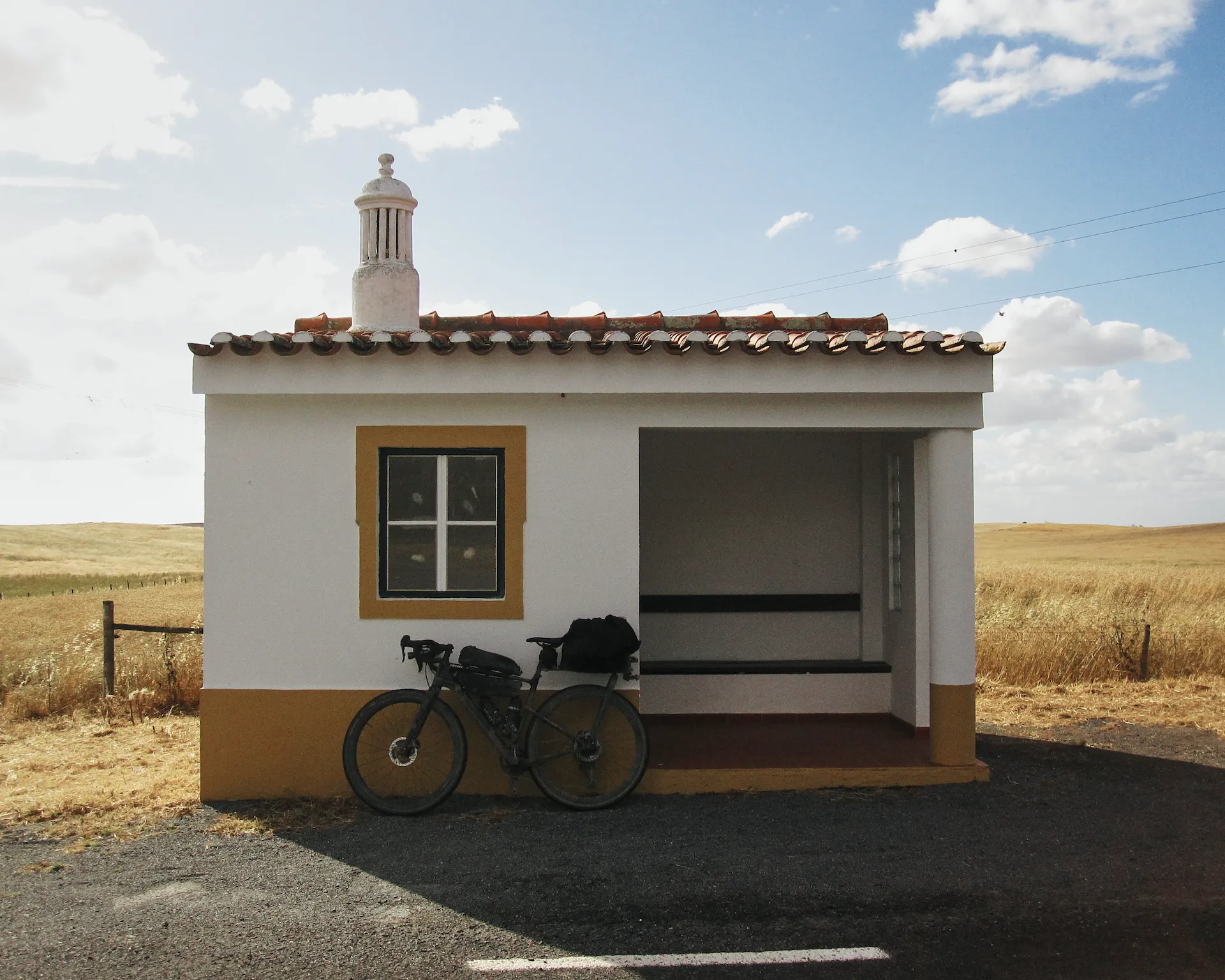 Loaded gravel bike leaning against a small white and ochre roadside shelter with a terracotta tile roof and traditional chimney, surrounded by golden wheat fields.