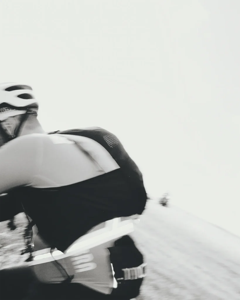 Blurred black and white close-up of a cyclist in a helmet and jersey riding fast, the bright empty background emphasising speed.