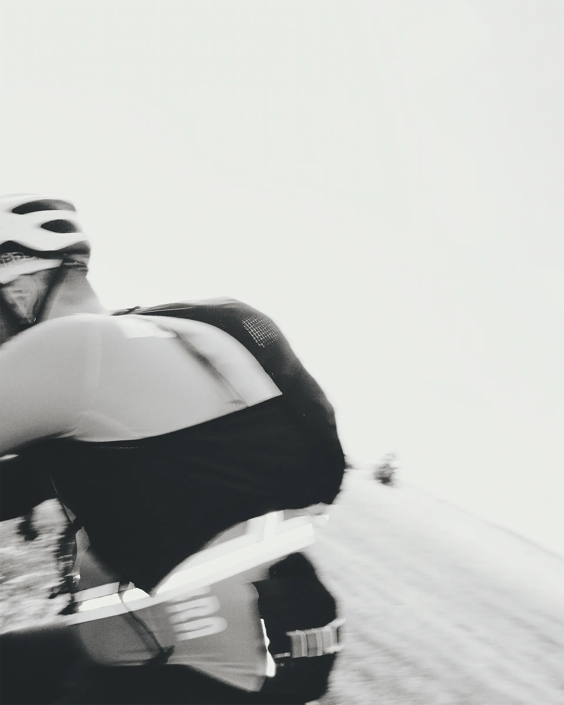 Blurred black and white close-up of a cyclist in a helmet and jersey riding fast, the bright empty background emphasising speed.