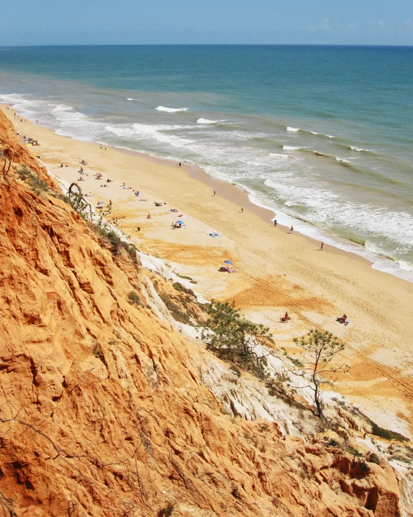 View down orange sandstone cliffs to a long golden Algarve beach with scattered sunbathers and waves rolling in from a turquoise ocean.