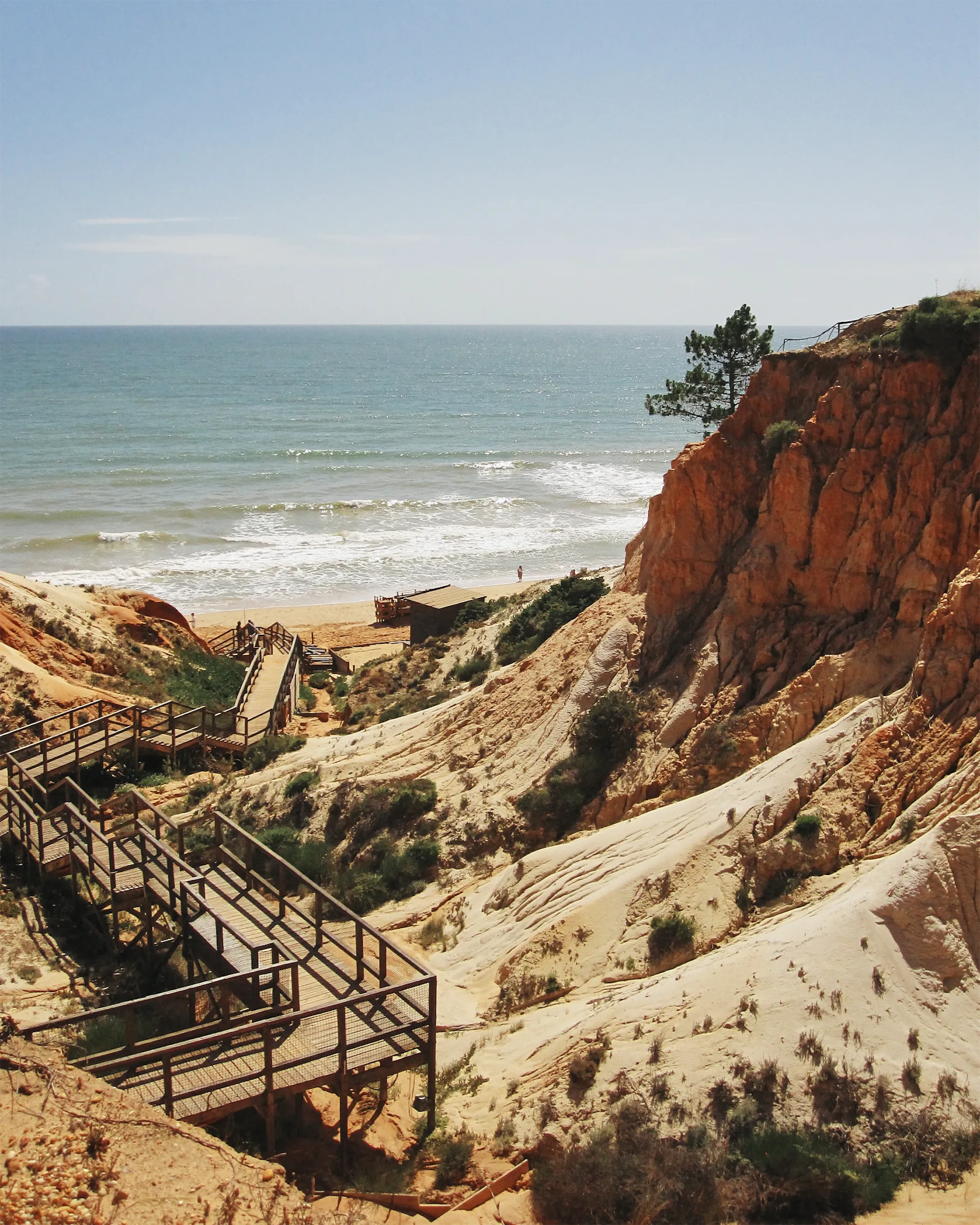 Wooden boardwalk stairs zigzagging down through eroded red and cream cliffs to a sandy beach and the Atlantic Ocean on a sunny day.