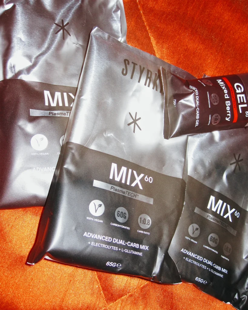 STYRKR MIX60 dual carb drink sachets and a GEL50 Mixed Berry energy gel laid out on an orange fabric background.